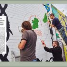 Texte in Bildern: "Belfaster Euphemismen: »Peace Line«, »Peace Wall«." Drei Personen malen ein Wandbild auf Ziegelwand.