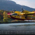 Kalender mit Daten unten, gelbes Löschflugzeug über Wasser, Bäume im Hintergrund.