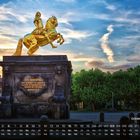 Ein goldener Reiter auf einem Pferd steht auf einem Sockel mit der Inschrift "FRID. AUGUSTUS I." Vor einem bewölkten Himmel.