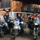 Drei Personen in Motorradkleidung sitzen auf Motorrädern und zeigen Daumen hoch, vor einem rustikalen Holzhaus.