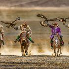 Drei Reiter in traditionellen Gewändern halten Adler auf erhobenen Armen, während sie über eine weite Ebene galoppieren.