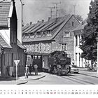 Text: „Mai“. Schwarz-Weiß-Szene: Eine Dampflokomotive fährt durch eine belebte Stadtstraße.