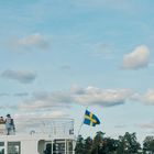 Ein weißes Boot mit schwedischer Flagge. Im Hintergrund Wasser, Bäume und ein Haus. Der Himmel ist leicht bewölkt.