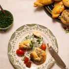 Ein Teller mit Grünmuster, darauf ein Brötchen mit Mohn, zwei Kirschtomaten und Schnittlauch. Ein Messer und ein gefüllter Teller daneben.