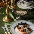 Ein dekorativer Teller mit Lachs, violetten Kartoffeln und Soße; daneben elegantes Besteck und ein prunkvoller Kerzenleuchter.