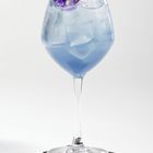 Ein hellblauer Cocktail mit Eis in einem Weinglas, garniert mit einer lila-gelben Blume, steht auf einem schwarzen Untersetzer.