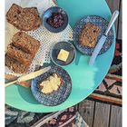 Drei Brotscheiben, Butter und Marmelade auf einem Tisch mit Teller und Messer. Eine gemusterte Decke ist darunter.