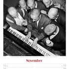 November. Eine Gruppe von Männern mit rasierten Köpfen steht um ein Klavier. Einer hält ein Getränk.