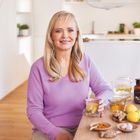 Eine Frau in einem lila Pullover sitzt an einem Tisch mit Ingwer, Zitrone und Honig.