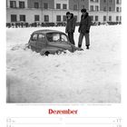 Dezemberkalender mit Bild zweier Männer und teils im Schnee vergrabenem Auto vor einem mehrstöckigen Gebäude.