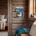 Kalender "Abenteuer Island" hängt an einer Holzwand in einem gemütlichen Raum mit Bett und dekorativen Vasen.