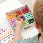Box mit bunten Holzkugeln, ein Kind sortiert sie mit einer Zange. Daneben eine Vorlage mit farbigen Punkten.