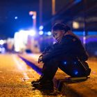 Eine Person in Uniform sitzt nachts auf dem Bordstein, Straßenlichter und ein unscharfes Fahrzeug im Hintergrund.