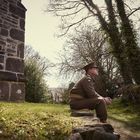 Ein Soldat in brauner Uniform sitzt draußen auf einer Steinmauer, neben einem alten Gebäude mit grauer Steinmauer.