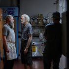 Oben links "PARTY ICE". Zwei ältere Männer stehen einander zugewandt in einem Lagerraum mit Kartons. Ein dritter Mann lehnt links.