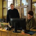 Es gibt einen „Confidential Waste“-Behälter. Ein Mann steht, eine Frau sitzt konzentriert vor einem Computer in einem Büro.