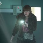 Eine Frau hält ein leuchtendes Handy in einem dunklen Raum, beleuchtet von einer Operationslampe; sie wirkt aufmerksam.