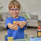 Ein Junge mit blauer Brille und Shirt hält Modelliermasse, lächelt fröhlich. Im Hintergrund wohnliches Zimmer.