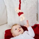 Ein Baby in weißer Kleidung liegt auf rotem Stoff und greift nach einem aufgehängten Plüschbären.