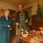 Ich sehe keine Texte. Zwei ältere Personen in einer Küche, eine Person zerkleinert Kräuter mit einem Fleischwolf.