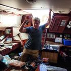 Kein Text sichtbar. Ein älterer Mann steht knietief in Wasser in einer überfluteten, unordentlichen Schiffs-Kajüte.