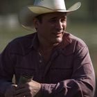 Ein Mann mit einem hellen Cowboyhut und lila Hemd lehnt entspannt an einem Holzzaun.
