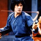 Ein Mann in einer blauen Robe hält Kampfstöcke, im Hintergrund zwei Mönche.