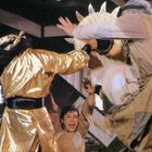 Person in einer goldenen Robe springt, während eine andere Person mit Schreien die Arme hebt, in einem Kampfsetting.