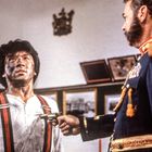 Ein Mann in Uniform hält eine Pistole auf einen erschöpften, schmutzigen Mann mit Hosenträgern. Im Hintergrund Bilder an der Wand.