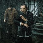 Ein Mann in traditioneller Kleidung steht im Regen, umgeben von anderen Männern und einer Treppe im Hintergrund.