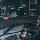 Ein Mann in grüner Jacke zielt mit einer Pistole auf den Fahrer eines Schnellboots in urbaner Umgebung.