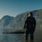 Text: "POLITI" auf der Jacke einer Person im Wasser, umgeben von Bergen.