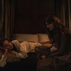 Zwei Frauen in einem dunklen Raum, eine liegt im Bett, die andere sitzt. Zwei Kerzen beleuchten die Szene sanft.