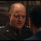 Ein Mann in Militäruniform mit „U.S.“-Abzeichen spricht ernst mit einer anderen Person in einem Innenraum.