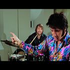 Keine sichtbaren Texte. Ein Mann in einem bunten Hemd zeigt mit dem Finger; im Hintergrund sind Musiker an Schlagzeug und Gitarre.