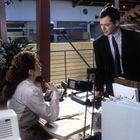 Zwei Personen in einem Büro, eine sitzend am Schreibtisch mit Telefon, die andere steht daneben, beide in Gespräch vertieft.
