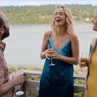 Drei Personen lachen und unterhalten sich auf einem Balkon mit Seeblick. Sie halten Sektgläser in den Händen.