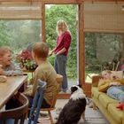 Ein heller Raum mit großen Fenstern, drei Kinder und ein Hund; eine Frau steht draußen und lächelt herein.