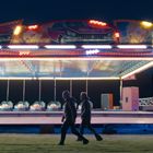 Beleuchteter Autoscooter auf einer Kirmes bei Nacht, mit bemalten Wänden und Lichtern. Zwei Personen gehen vorbei.
