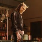 Ein bedrohlich wirkender Maskierter in einer Küche mit Töpfen, Mehlfäden auf Kleidung und Axt in der Hand.