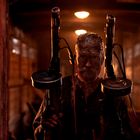 Ein Mann mit schmutzigem Gesicht steht in einem dunklen, holzverkleideten Raum und hält zwei Maschinengewehre.