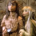 Zwei Puppenwesen mit spitzen Ohren und langen Haaren stehen vor einer geschnitzten Wand. Ein Wesen hält einen Kristall.