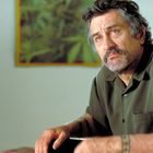 Ein nachdenklich wirkender Mann in einem olivgrünen Hemd mit Bart, Tattoos an Armen und sommerlich hellem Hintergrund.