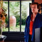 Ein Mann in blauem Hemd steht neben einem Blumenarrangement vor einem großen Fensterausblick ins Grüne.