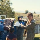 Ein Mann zeigt auf einen blauen Traktor. Ein Kind steht daneben und schaut zu. Ländliche Landschaft im Hintergrund.