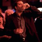 Eine Person im Anzug trinkt an der Bar, umgeben von einer geselligen Gruppe im gedämpften Licht.