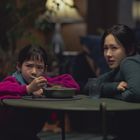 Ein Mädchen in pinkem Pullover sitzt mit Löffel vor einer Schale, daneben eine Frau in grünem Pullover am Tisch.