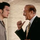 Ein junger Mann in Arbeitskleidung und ein älterer Mann im Anzug diskutieren lebhaft in einem hellen Raum.