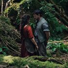 Im dichten Wald stehen ein Mann und eine Frau einander nahe gegenüber, umgeben von Moos und Wurzeln.