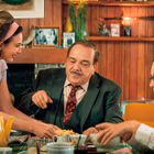 Eine Frau, ein Mann und ein Junge sitzen an einem gedeckten Tisch. Der Raum hat eine gemütliche, retro Atmosphäre.